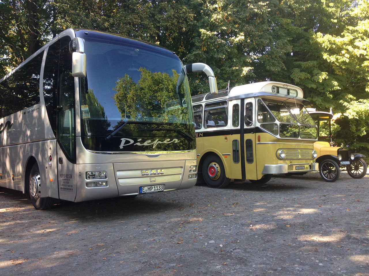 Drei Reisebusse aus unterschiedlichen Zeitepochen stehen nebeneinander auf einem Parkplatz unter Bäumen in der Sonne